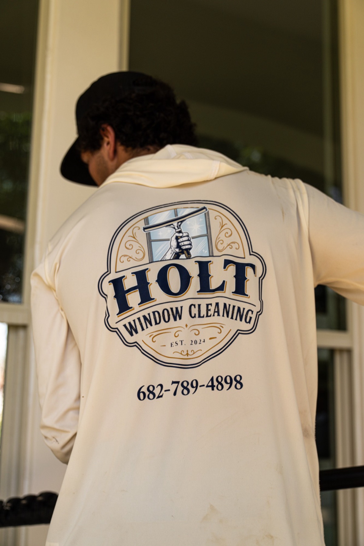 Caleb Holt at a customer's front windows, the Holt Window Cleaning logo across the back of his shirt.